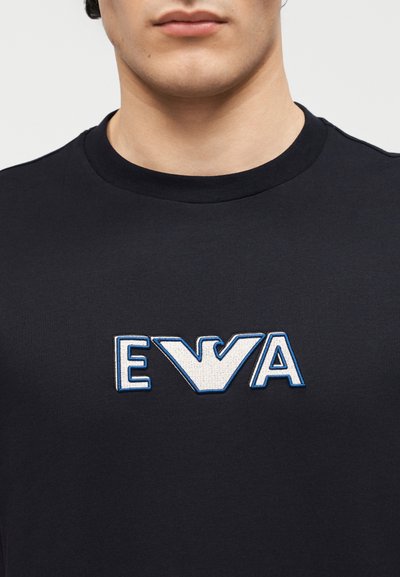 Κοντινό πλάνο ενός ατόμου που φοράει μαύρο πουκάμισο με ένα λευκό και μπλε κεντημένο λογότυπο που συνδυάζει τα γράμματα E, ένα στιλιζαρισμένο πουλί και το γράμμα A στο στήθος.