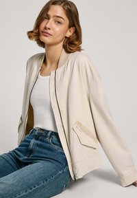 Veste bomber beige clair avec une texture lisse, col côtelé, fermeture éclair et deux poches avant, portée par-dessus un débardeur blanc et un jean bleu.