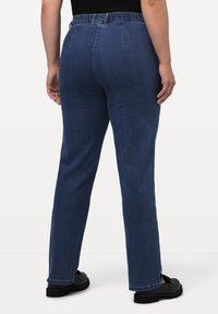 Pantalones de mezclilla de color azul oscuro, con forma de pierna recta, cinturilla elástica y detalles de costura mínimos. Llevados con zapatos negros.