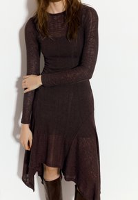 Langærmet, ribbestrikket kjole i mørkebrun med en flagrende kant. Har en tætsiddende silhuet og tekstureret stof.