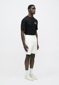 Vīrietis stāv, valkājot melnu T-kreklu ar EA7 logotipu, baltas elastīgas vidukļa šortus, baltas zeķes un baltus sporta apavus uz vienkrāsaina gaiša fona.