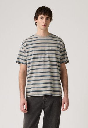 Jonge man met een beige en blauw gestreept T-shirt met korte mouwen en donkergrijze broek, staand tegen een effen achtergrond.