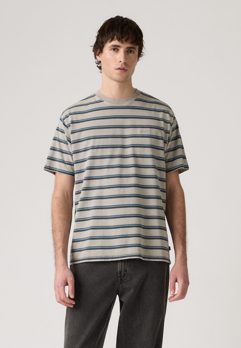 Young man wearing a beige and blue striped short-sleeve T-shirt and dark gray pants, standing against a plain background.