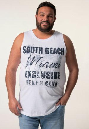 Biała koszulka na ramiączkach z czarnym nadrukiem tekstu "SOUTH BEACH Miami EXCLUSIVE BEACH CLUB" oraz wzorem palm; zestawiona z jasnoniebieskimi dżinsami.