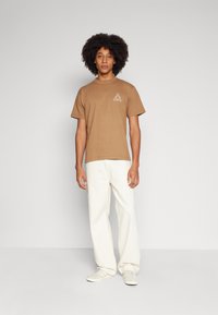 Camiseta de algodón marrón con mangas cortas y un gráfico en forma de triángulo en el lado izquierdo del pecho, combinada con pantalones de pierna ancha color blanco sucio y zapatillas ligeras.