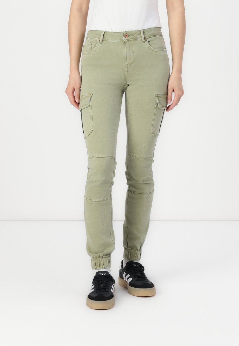 Personne portant un pantalon cargo vert clair avec des poignets élastiques et des baskets noires à rayures blanches, debout devant un fond blanc.