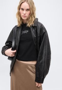 Jeune femme portant une veste en cuir noire, un crop top noir avec le texte "Juice", une jupe beige et un sac à bandoulière noir sur un fond uni.