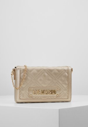 Sac à main Love Moschino matelassé doré avec bandoulière en chaîne et plaque logo en métal, exposé sur une surface blanche contre un fond uni.