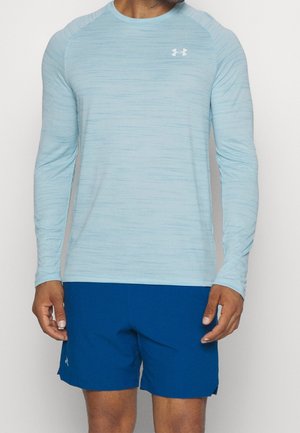 Mann trägt hellblaues langärmliges Sportshirt und dunkelblaue Shorts, steht vor einem einfarbigen Hintergrund.