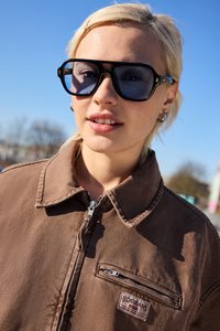 Mladý človek s krátkymi blond vlasmi nosí veľké tmavé slnečné okuliare a hnedú bundu na zips vonku na jasný, slnečný deň.