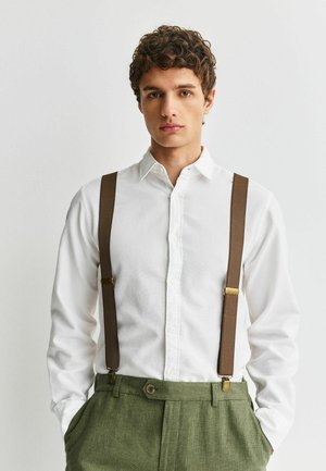 Young man with curly hair wearing a white button-up shirt, brown suspenders, and green textured trousers, hands in pockets, neutral expression.