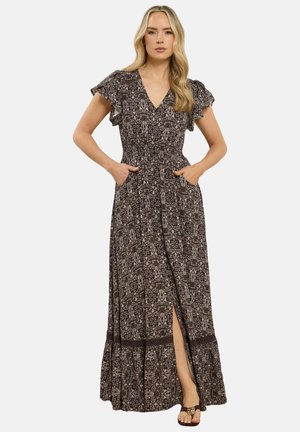 Vrouw met lang blond haar die een bruine maxi-jurk met patroon draagt, met wappermouwen, een split aan de zijkant en handen in de zakken, staand tegen een witte achtergrond.