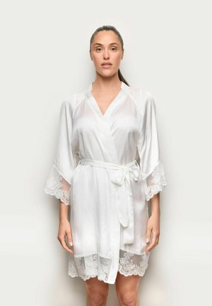 Frau mit zurückgebundenem dunklem Haar, die ein durchsichtiges weißes Morgenmantel mit Spitzenbesatz und einem Taillengürtel trägt, vor einem schlichten hellen Hintergrund stehend.