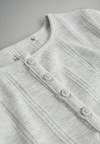 Haut côtelé gris clair présentant un col arrondi et six boutons blancs sur le devant, confectionné en coton doux avec une finition texturée.