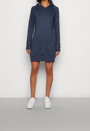 Woman wearing a navy blue hoodie dress with front pockets and white sneakers, standing against a plain light background.