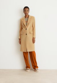 Cappotto di lana beige con un design su misura, rever a forma di notched e due tasche frontali. Abbinato a pantaloni color ruggine e sneaker casual.