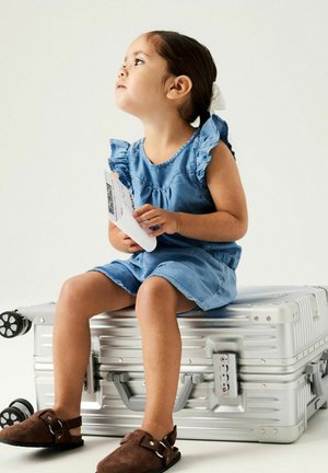 Młoda dziewczyna w niebieskiej sukience trzyma kartę pokładową, siedzi na stosie srebrnych walizek, patrząc w górę na białym tle.