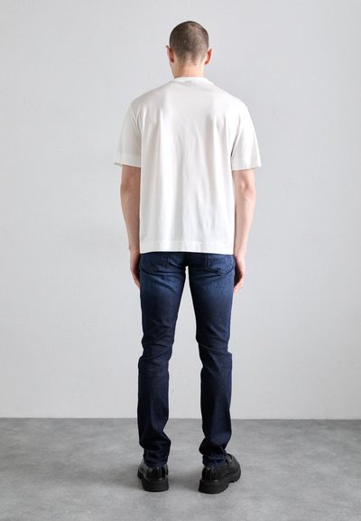 Weißes Baumwoll-T-Shirt mit kurzen Ärmeln, kombiniert mit dunkelblauen Jeans. Das Outfit wird mit schwarzen flachen Schuhen abgerundet. Rückansicht.