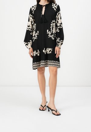 Femme portant une robe noire longueur genou avec des motifs floraux blancs, manches longues, col avec lien à pompons, et des sandales noires à talons avec fines lanières.