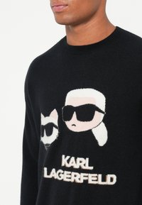 Fekete kötött pulóver, amelyen egy macska és napszemüveget viselő személy karikatúrája látható, alatta pedig "KARL LAGERFELD" felirat krém színben.