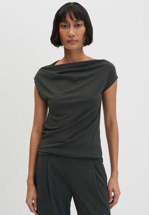 Vrouw met kort donker haar, gekleed in een zwarte mouwloze gedrapeerde top en bijpassende zwarte broek tegen een effen lichte achtergrond.