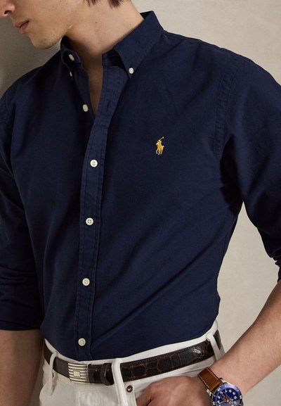 Vyras dėvi tamsiai mėlynį marškinius su sagutėmis ir geltonu logotipu, baltas kelnes, rudos odos diržą ir mėlyną riešo laikrodį su rudos odos dirželiu.