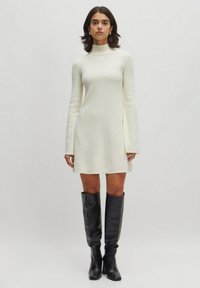 Knee-langes cremefarbenes Kleid mit langen Ärmeln und einem hohen Kragen, kombiniert mit kniehoche schwarzen Lederstiefeln. Glatte Textur, minimales Design.