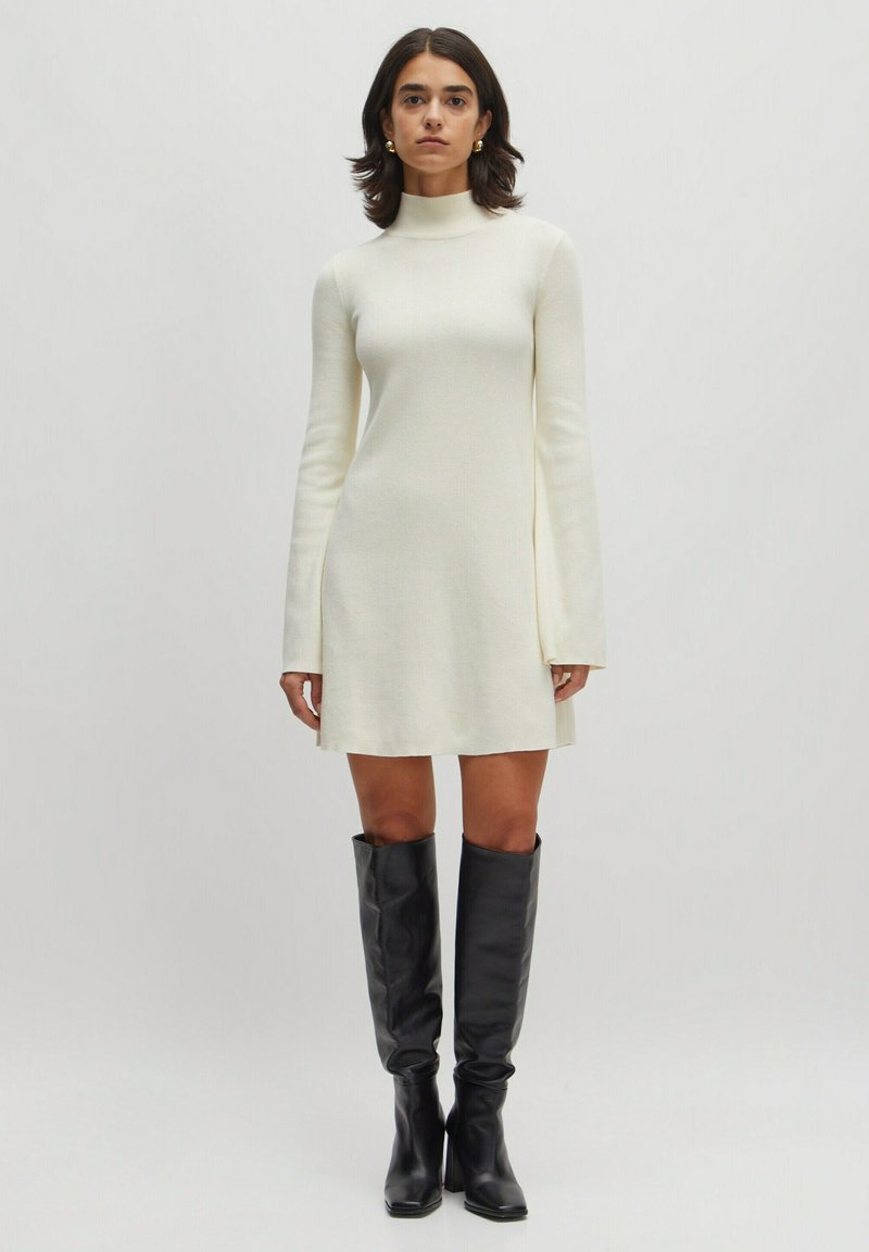 Knee-langes cremefarbenes Kleid mit langen Ärmeln und einem hohen Kragen, kombiniert mit kniehoche schwarzen Lederstiefeln. Glatte Textur, minimales Design.