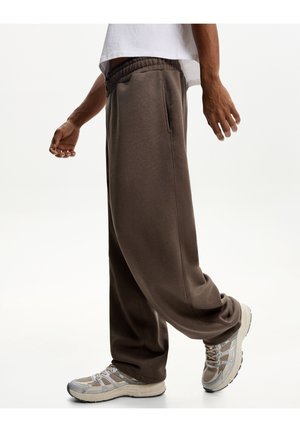 Braune Baumwoll-Sweatpants mit relaxter Passform, ausgestattet mit einem elastischen Bund und seitlichen Taschen, kombiniert mit beige-farbenen Sportschuhen.