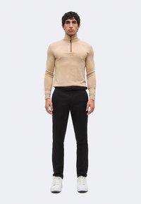 Homme debout de face portant un pull zippé beige, un pantalon noir avec ceinture et des baskets blanches sur un fond uni.