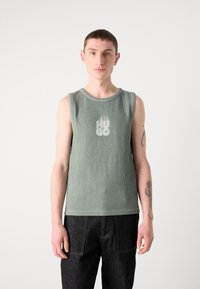 Zelený rebrovaný bezrukávový top s okrúhlym výstrihom a vyblednutým grafickým logom na hrudi, kombinovaný s tmavými džínami.