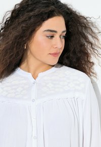 Camicia bianca con bottone, decorata con un motivo floreale ricamato sulla spalla, in tessuto leggero, design plissettato e colletto rotondo.