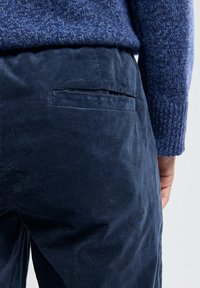 Pantalons en velours côtelé bleu marine avec une texture lisse et une poche arrière. Le tissu semble légèrement usé, associé à un pull en maille bleu.