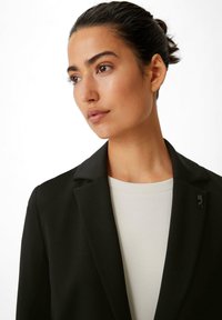 Schwarzer Blazer mit glatter Textur, ausgestattet mit Reverskragen und einem schwarzen Pinnadel-Akzent. Getragen über einem hellen, taillierten Oberteil.
