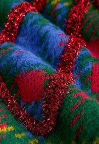 Pletivová látka s vzorom v červenej, zelenej a modrej farbe, zdobená lesklým červeným trblietavo. Mäkka textura, vrstvený dizajn s kontrastnými farbami.