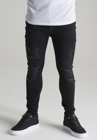 Zwarte skinny jeans met versleten scheuren op de knieën, gecombineerd met zwarte sneakers met witte zolen en gouden accenten.