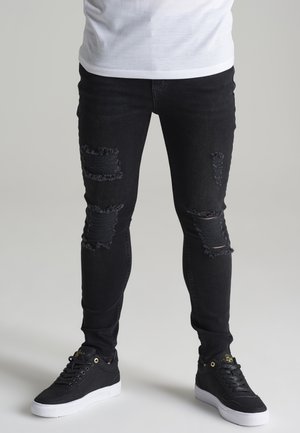 Jeans skinny noirs avec des déchirures au niveau des genoux, associés à des baskets noires avec des semelles blanches et des accents dorés.