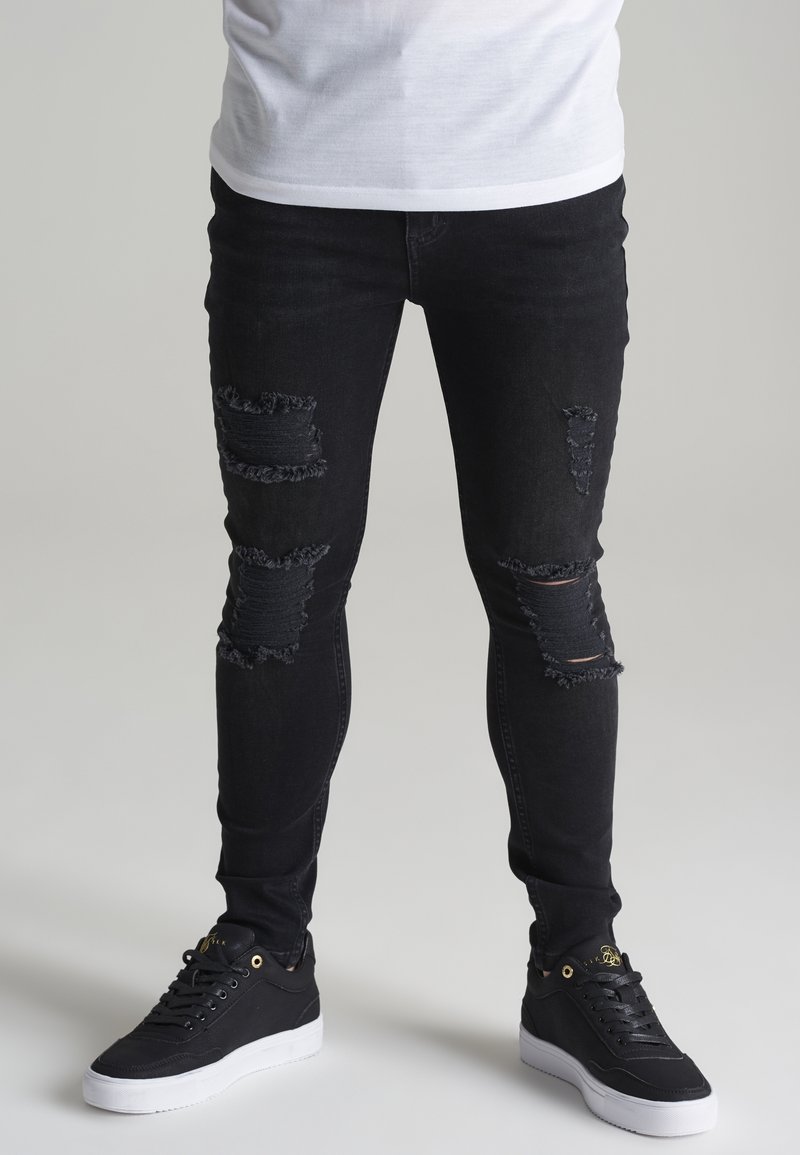 Zwarte skinny jeans met versleten scheuren op de knieën, gecombineerd met zwarte sneakers met witte zolen en gouden accenten.