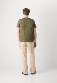 Quilted olive green vest over a brown t-shirt, paired with cream cargo pants. The vest features a simple, sleeveless design without hardware.