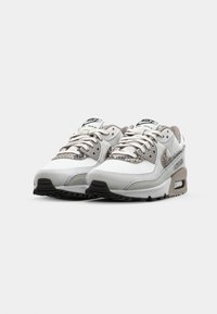 Balti un pelēki Nike Air Max sporta apavi ar teksturētām virsmām, auklu aizdari un redzamu gaisa spilvenu zole. Melna gumijas zole.
