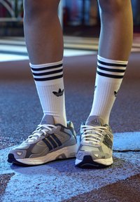 Sive i crne atletske cipele s bijelim vezicama; nose se s bijelim čarapama koje imaju tri crne pruge i crni logotip.