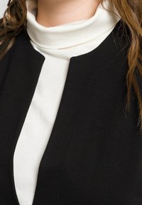 Svart kavaj med rund krage över en vit polotröja. Kavajen har en minimalistisk design och en slät textur.