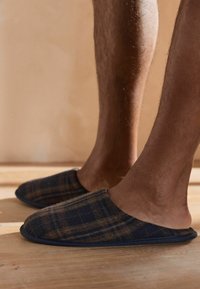 Rutede tøfler i mørkeblå og brun stof med slip-on design, runde tåspidser og en blød, struktureret overflade. Skridsikre såler synlige.
