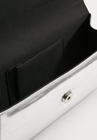 Liten silverfärgad clutchväska som är öppen och visar ett svart tyginteriör med en ficka och en metallisk tryckknappslås.