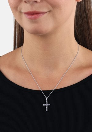 Colgante de cruz plateada en una cadena delgada, usado alrededor del cuello sobre una blusa negra de canalé, mostrando la parte inferior de la cara y las clavículas.