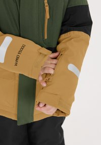 Buitenjas met groene, zwarte en tan secties. De tan mouwen hebben reflecterende accenten en een ritszak. Gemaakt van waterdicht materiaal.