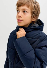 Jauns cilvēks ar īsiem gaiši brūniem matiem, ģērbies tumši zilos pufīgiem mētelī, turētu apkakli pie zoda, skatoties uz augšu.