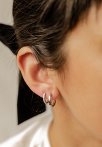 Srebrne kolczyki koła z fakturowanym wzorem, jeden ozdobiony małymi przezroczystymi kryształkami, zapięte na płatku ucha. Włosy związane do tyłu, neutralne tło.