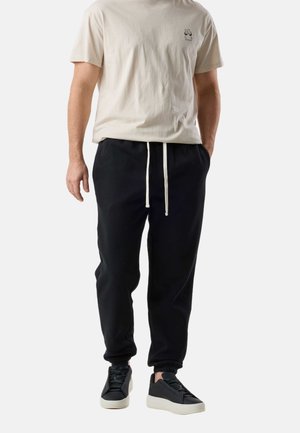 Homme portant un t-shirt beige à manches courtes avec un petit motif sur la poitrine, un pantalon de jogging noir avec cordon de serrage, et des baskets noires avec semelles blanches.