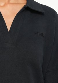 Černý pulovr s límečkem, s designem do V a texturovanou tkaninou. Malá černá výšivka loga na hrudi.